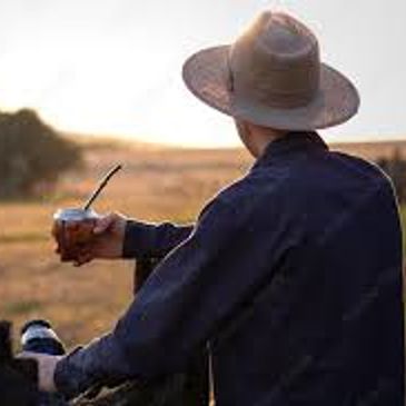 Man in a hat drinking mate, enjoying a sunset outdoors.