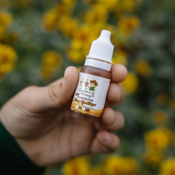 Hand holding a small bottle of Queen's Blessing honey drops with a blurred floral background.
