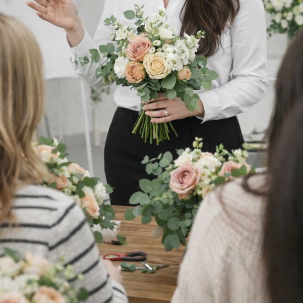 Woman teaching flower arranging to a group, holding a bridal bouquet.