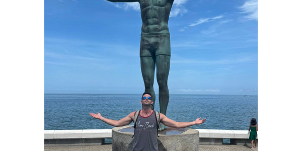 Man mimics statue's pose with arms outstretched by the ocean.