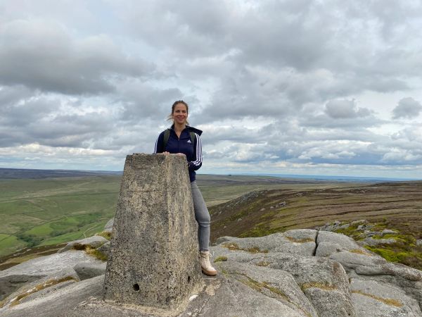 Woman standing beside a trig point on a rocky hilltop under a cloudy sky.