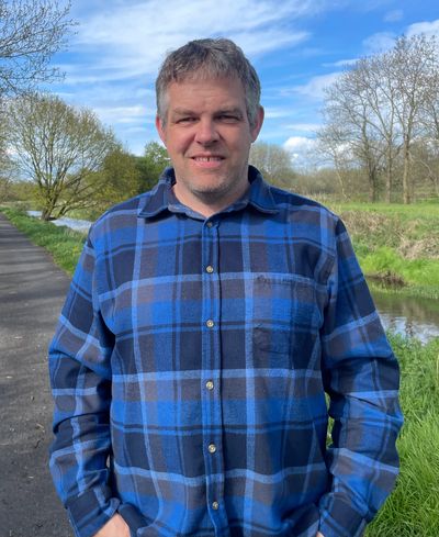 Man in a blue plaid shirt standing outdoors by a path and river.
