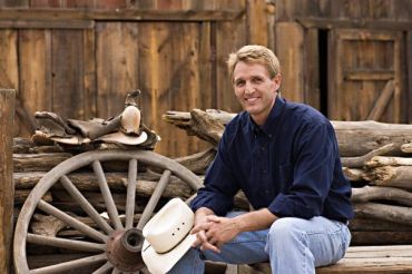 Man sitting near a wagon wheel holding a cowboy hat.