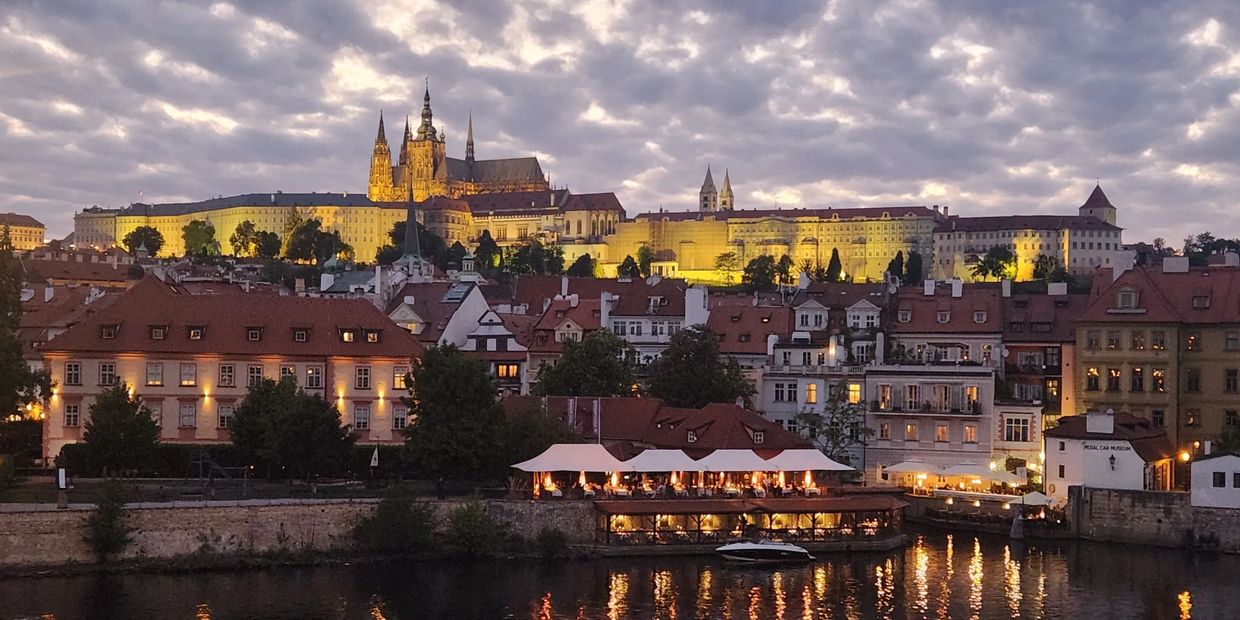 Evening view of illuminated Prague Castle and riverside buildings with reflections on the water.