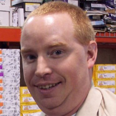 Man smiling in a warehouse with shelves of boxes behind him.