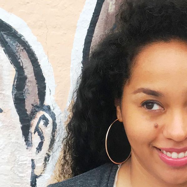Close-up of a smiling woman with curly hair and hoop earrings beside a wall with an ear painting.