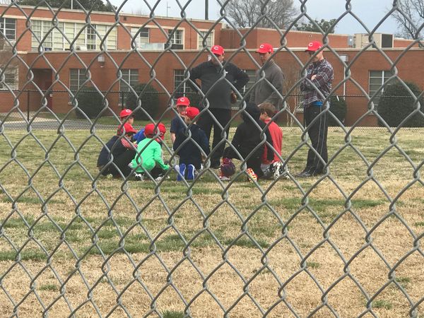 Youth baseball team in red caps listening to coaches behind a chain-link fence.