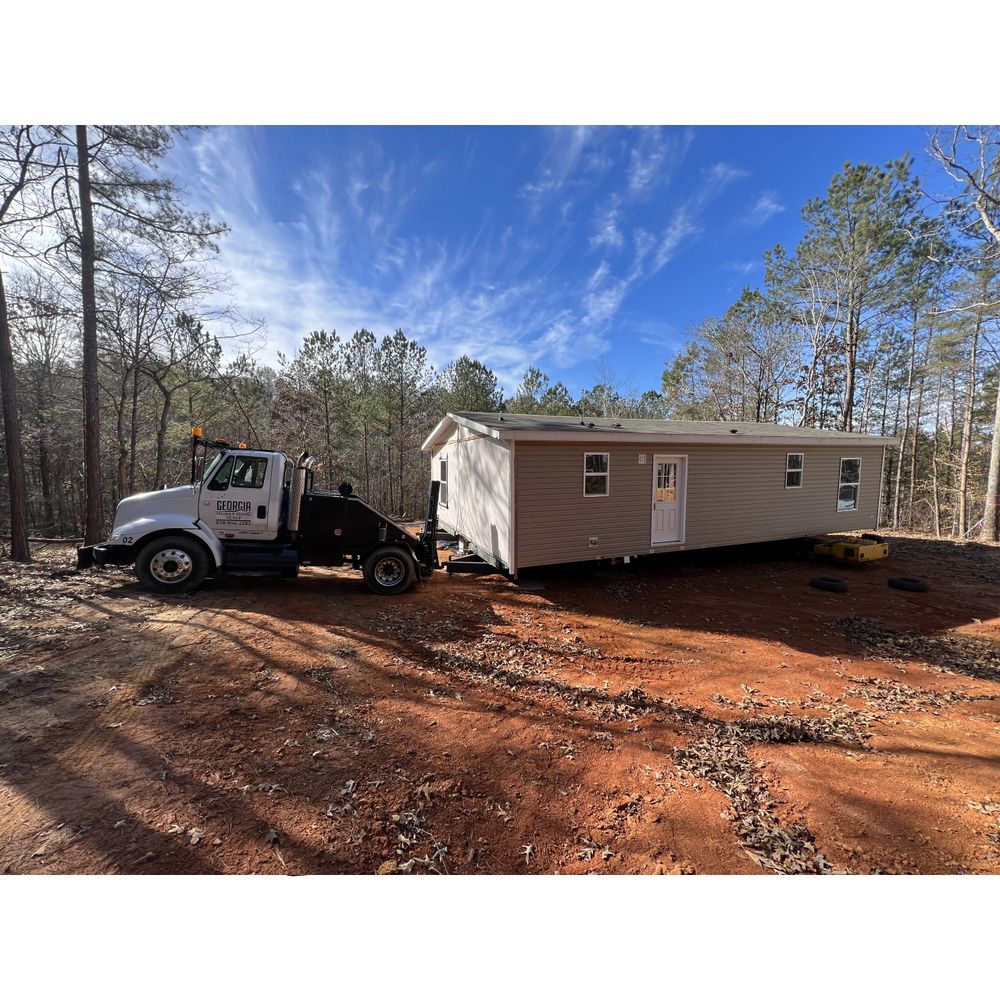 Truck transporting a manufactured home on a dirt clearing.