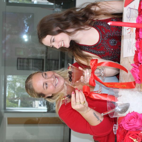 Two women cheer with cocktails decorated with red ribbons at a festive table.