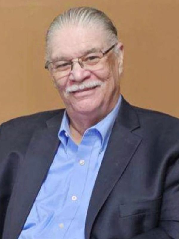 Smiling elderly man with glasses, wearing a blue shirt and dark blazer.