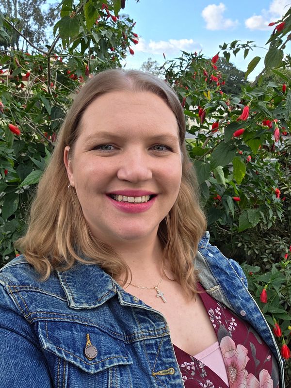 Smiling woman in denim jacket with green leafy background.