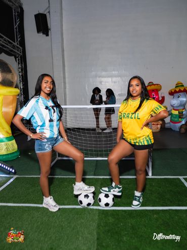 Two women in soccer jerseys posing with soccer balls on an indoor field.