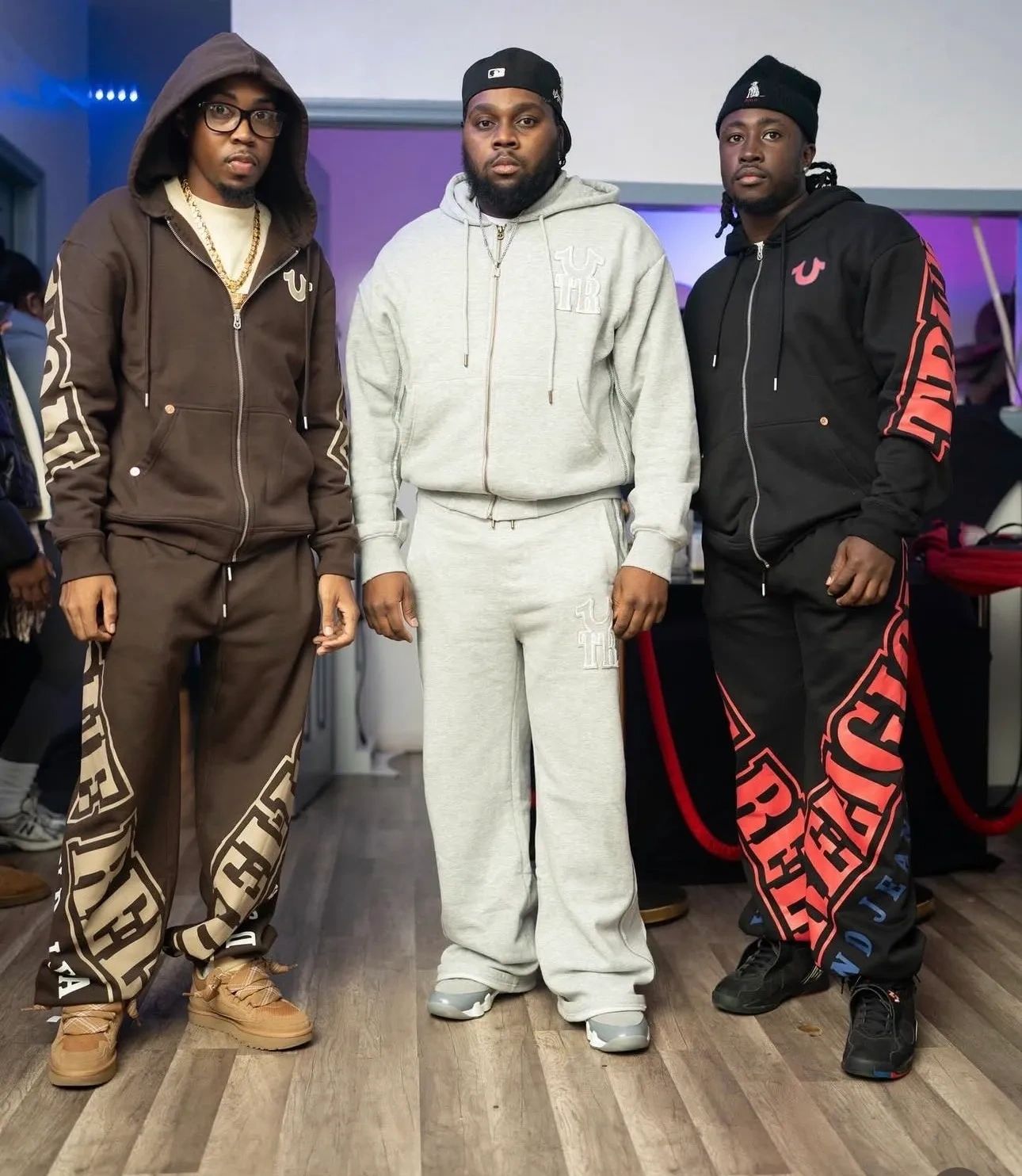 Three men wearing stylish hoodies and sweatpants standing indoors on a wooden floor.