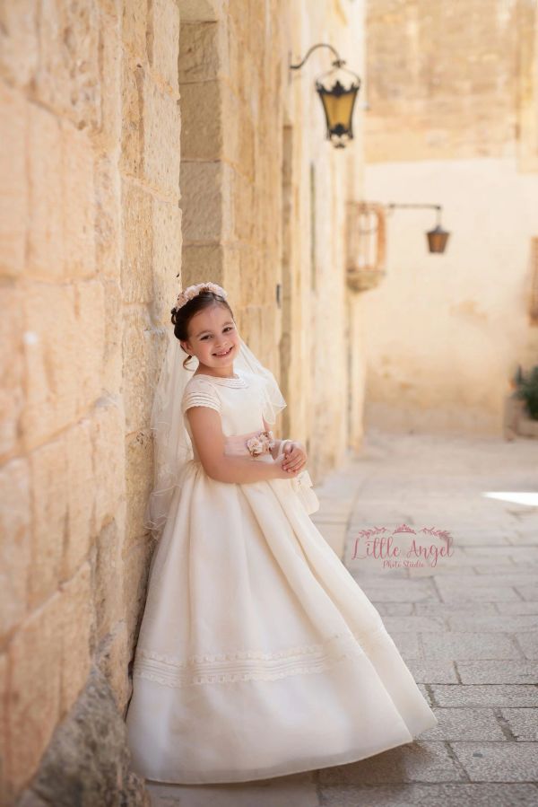 Smiling girl in a white dress with a veil, standing against a stone wall.