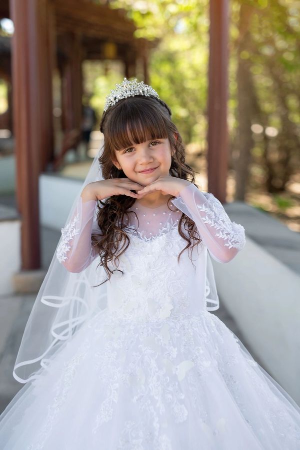A young girl in a white, ornate dress and tiara poses with a sweet smile outdoors.