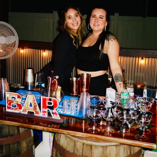 Two women smiling behind a well-stocked bar with cocktail glasses and mixers.