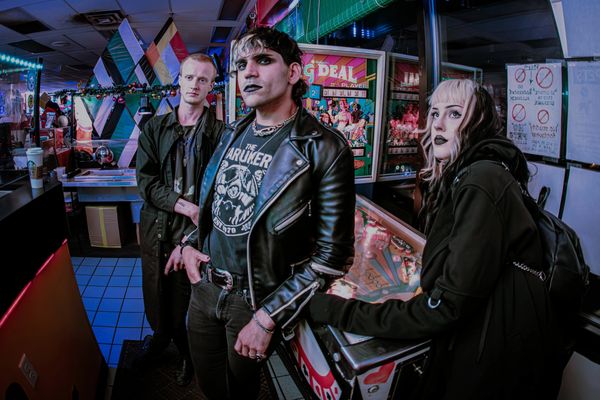 Three goth-style individuals pose in a neon-lit arcade near a pinball machine.