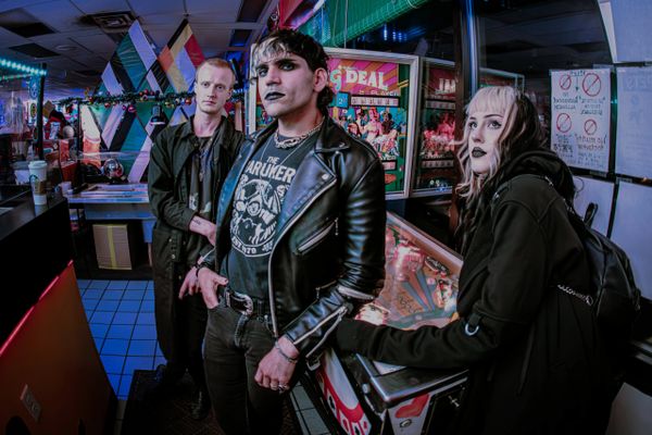 Three goth-style individuals pose in a neon-lit arcade near a pinball machine.