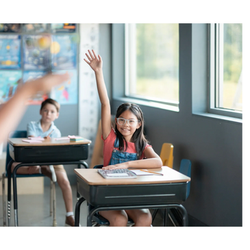 A young girl eagerly raises her hand in a bright classroom.