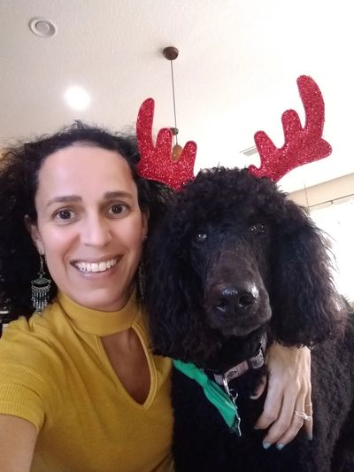 Waleska is standing next to her black poodle who is wearing a headband with reindeer antlers.