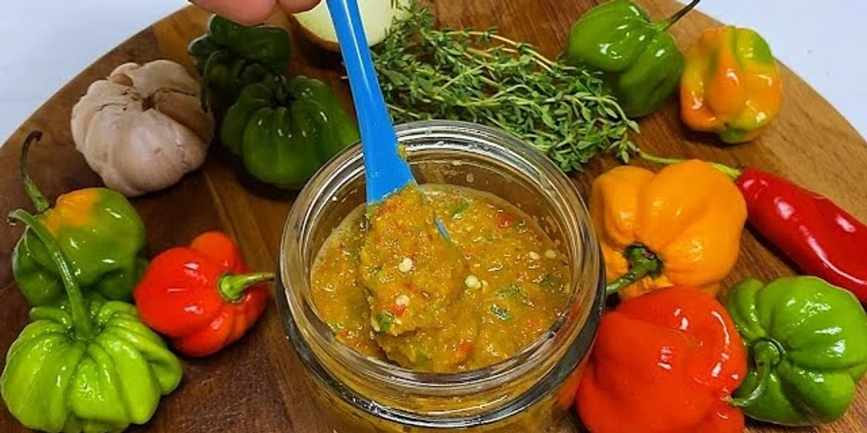 A jar of spicy homemade pepper sauce surrounded by fresh peppers and herbs on a wooden board.