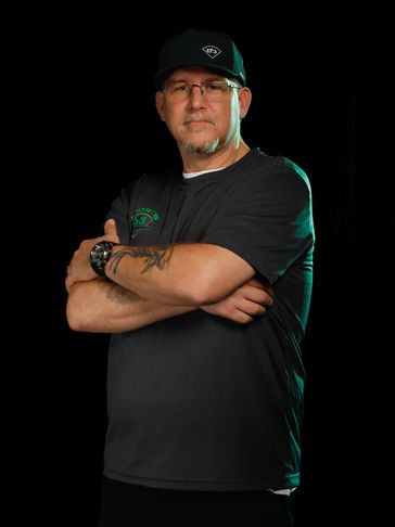 Man wearing glasses and a cap, arms crossed, with tattoos and a watch.