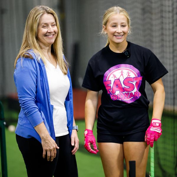 Two women smiling, one in sports gear holding a bat, the other in casual wear.