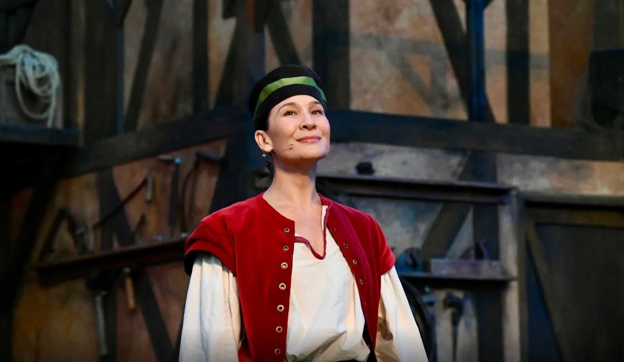 A woman in medieval costume performs on stage with a confident smile.