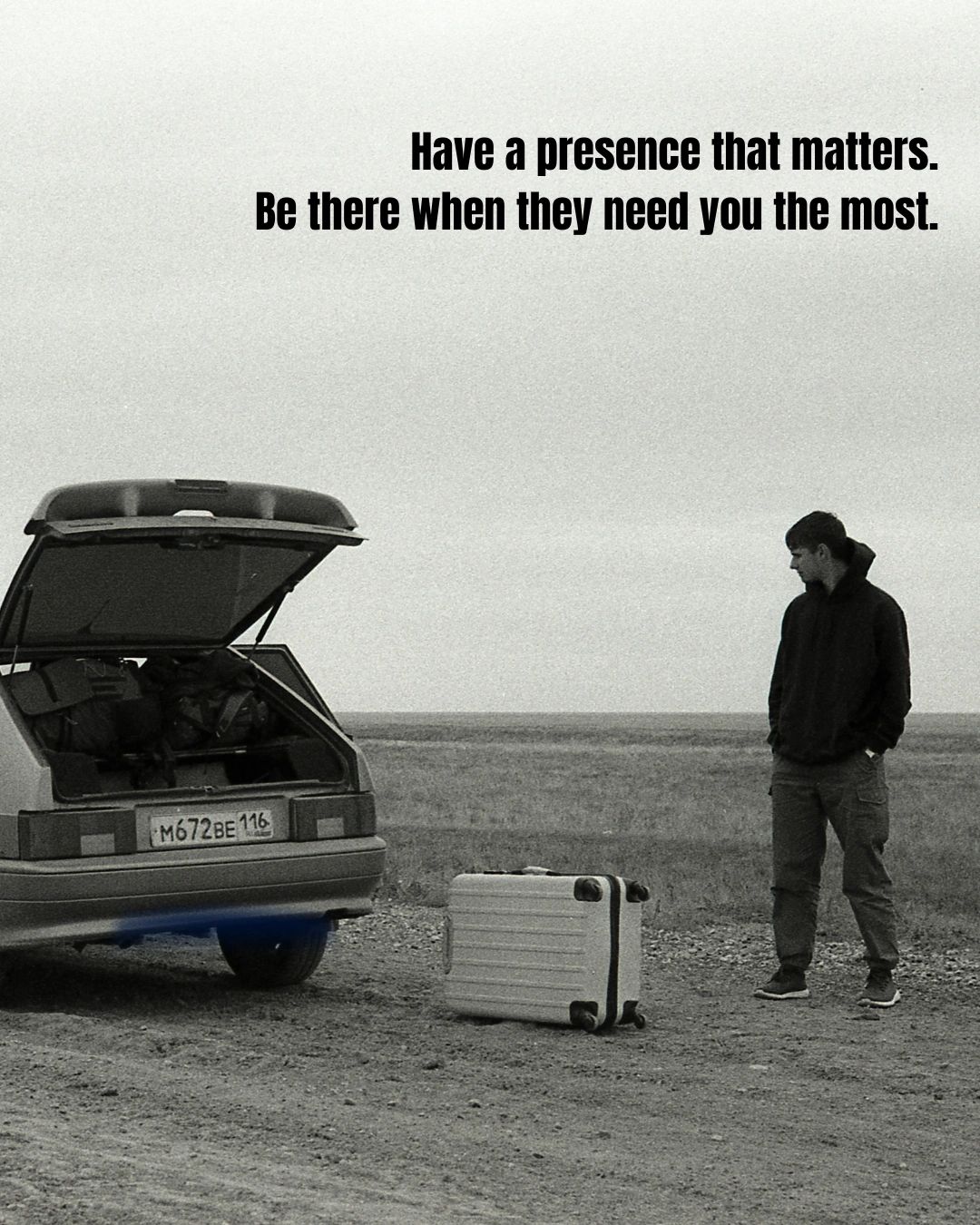 A man stands near an open car trunk and suitcase in a barren landscape.