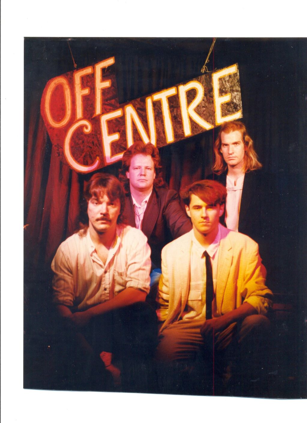 Four men posing under a glowing "OFF CENTRE" sign with a theatrical backdrop.