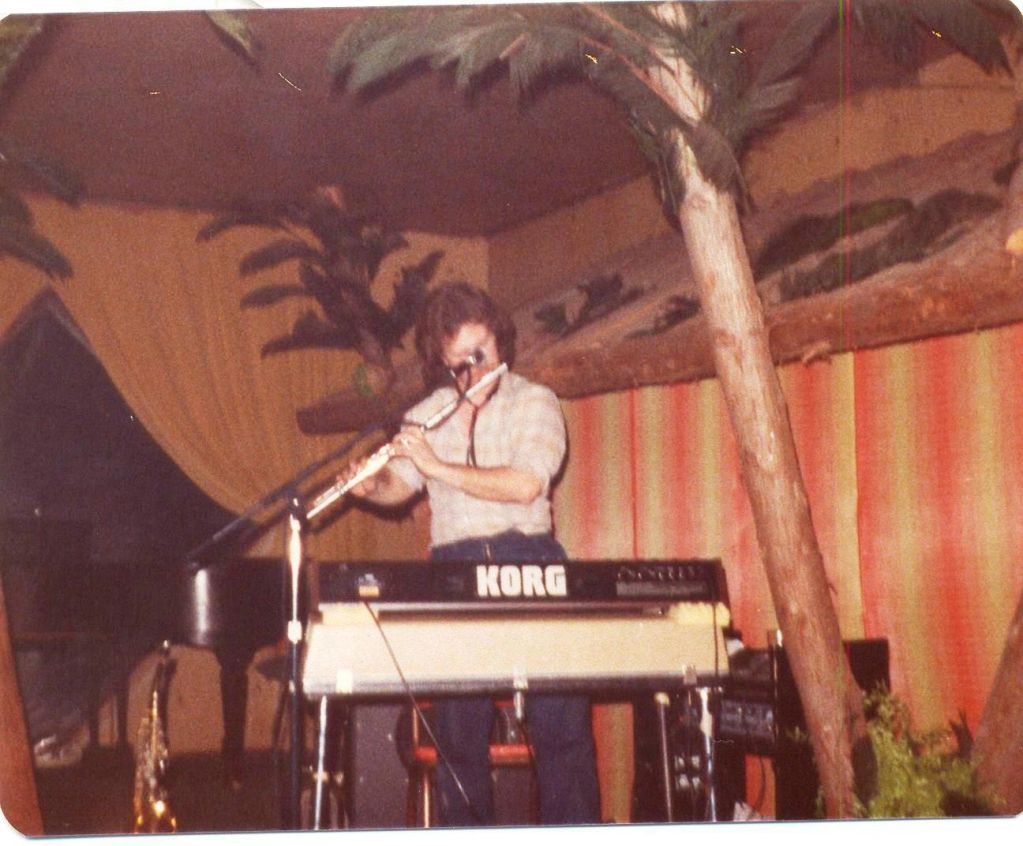 Musician playing flute behind a Korg keyboard in a retro room.