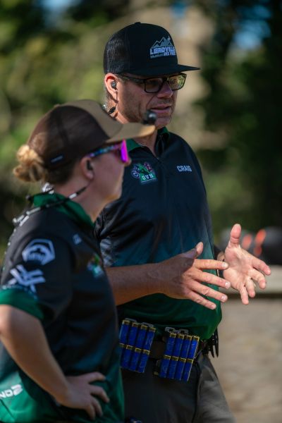 Two people in team uniforms discuss, one wearing a hat with shotgun shells.