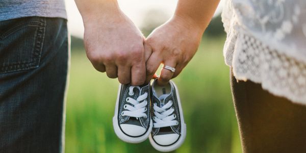 Closeup of the hands of a man and wife in a field linked together, holding the shoes of their child.