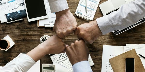 A business team fist-bumping over a table with calculator, smartphones, tablets, and important documents.