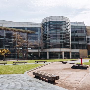 Modern glass building with a courtyard and benches under a cloudy sky.
