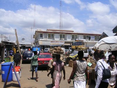 Busy market street with vendors carrying goods on their heads under a partly cloudy sky.