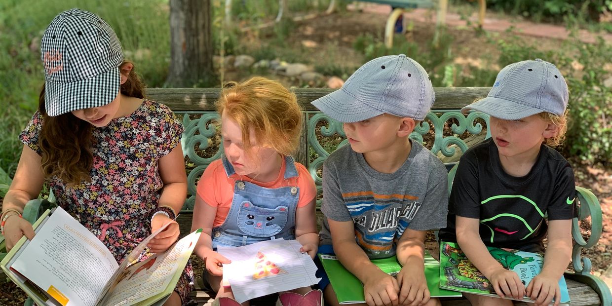 Four children sitting on a bench, reading books outdoors.