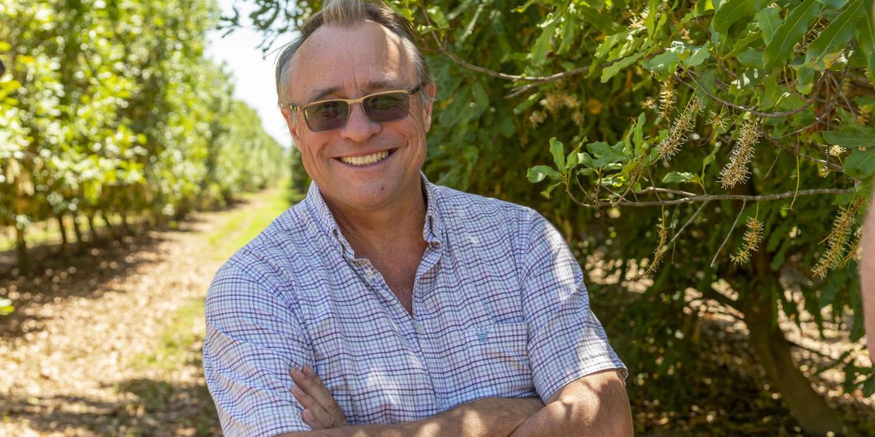 Smiling man with sunglasses stands in a sunny orchard with arms crossed.