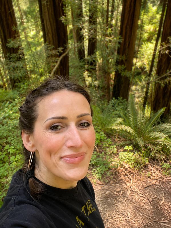 Woman smiling in a forest with sunlight filtering through trees.