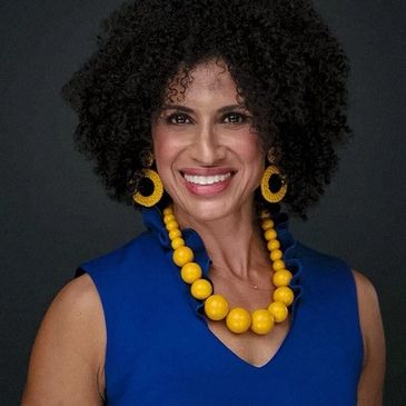 Smiling woman with curly hair wearing a blue dress and yellow accessories.