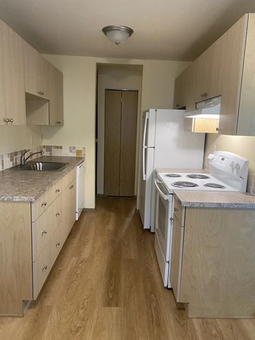 Narrow kitchen with wooden cabinets, white appliances, and wood flooring.