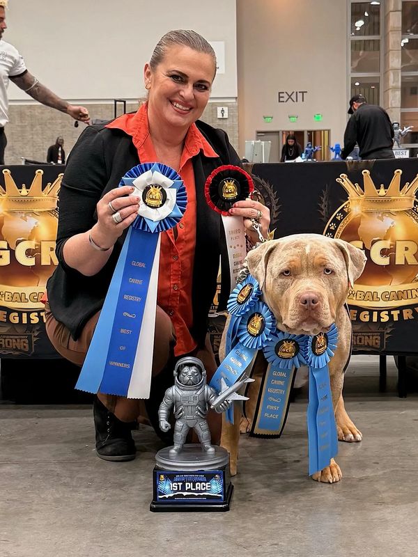 Woman and dog with multiple blue ribbons and a 1st place trophy at a dog show.