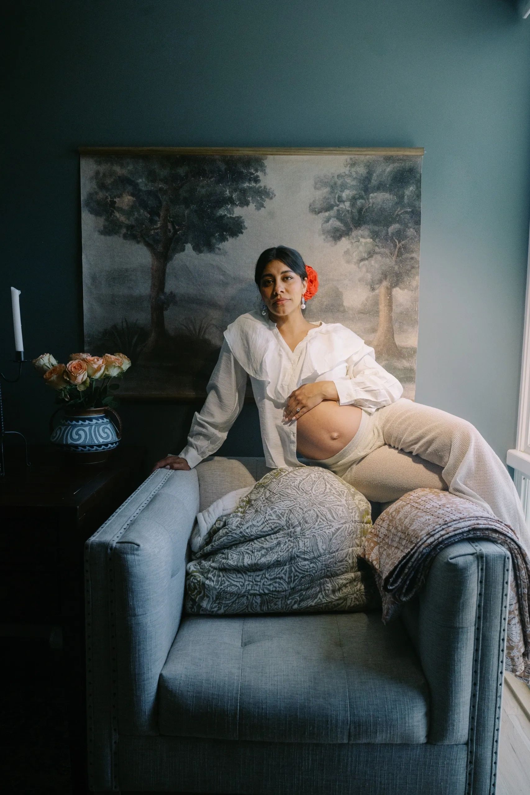 Pregnant woman with a red flower in her hair sits on a couch.