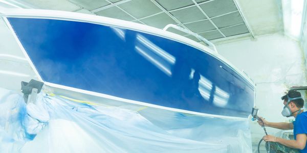 Person spray painting a boat's hull blue in a workshop.