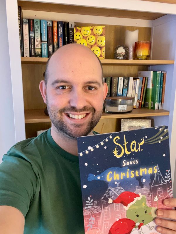 Smiling man holding a Christmas-themed book in front of a bookshelf.