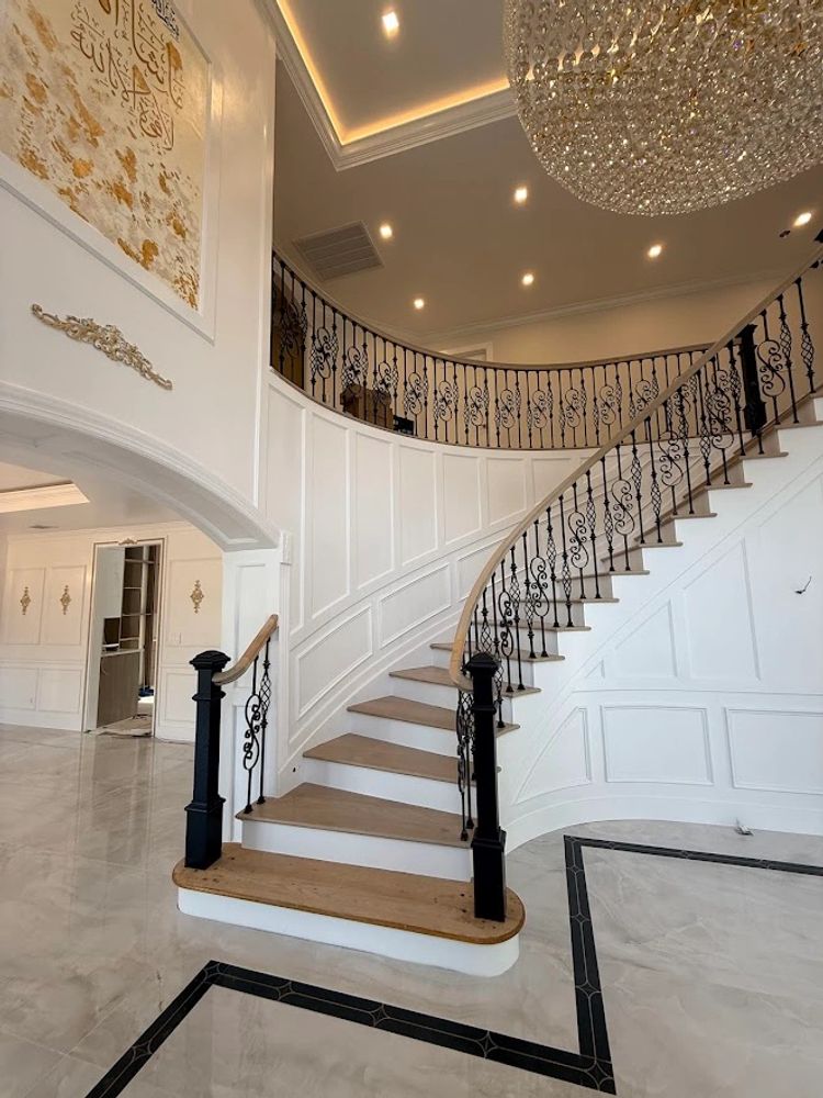 Elegant curved staircase with intricate black railings and a large chandelier in a luxurious white interior.