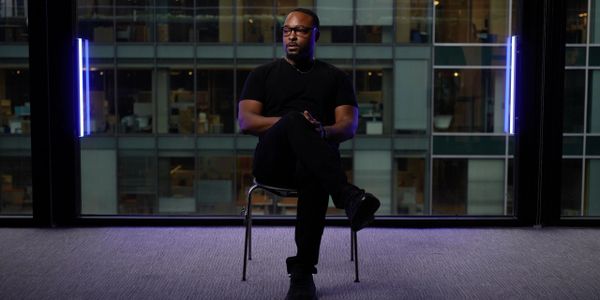 A man in black sits cross-legged on a chair in a modern office space.