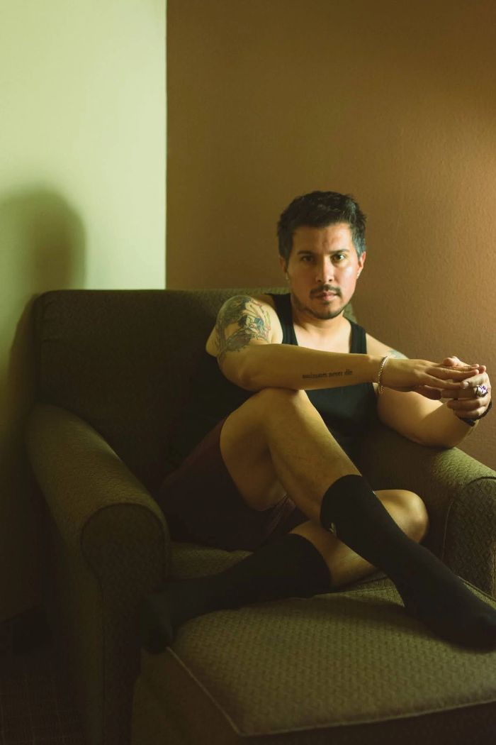 Man with tattoos sitting on a green armchair in a dimly lit room.