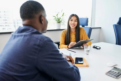 Two people, possibly in an interview, are seated at a desk, with one holding a clipboard