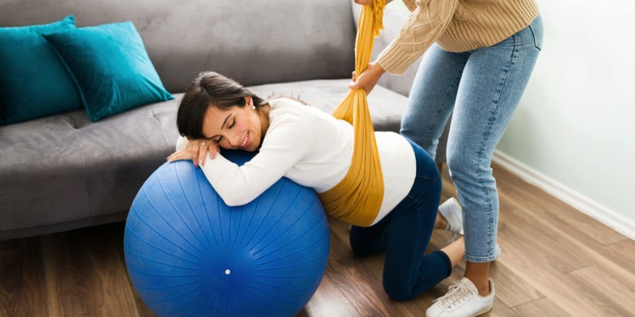 Pregnant woman receiving support with a birthing ball and sling at home.